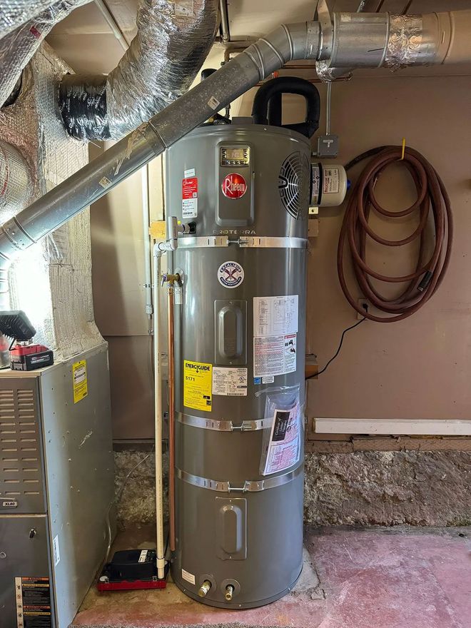 Tall, gray water heater in a basement setting. Metal ductwork and a reddish-brown water hose are visible.