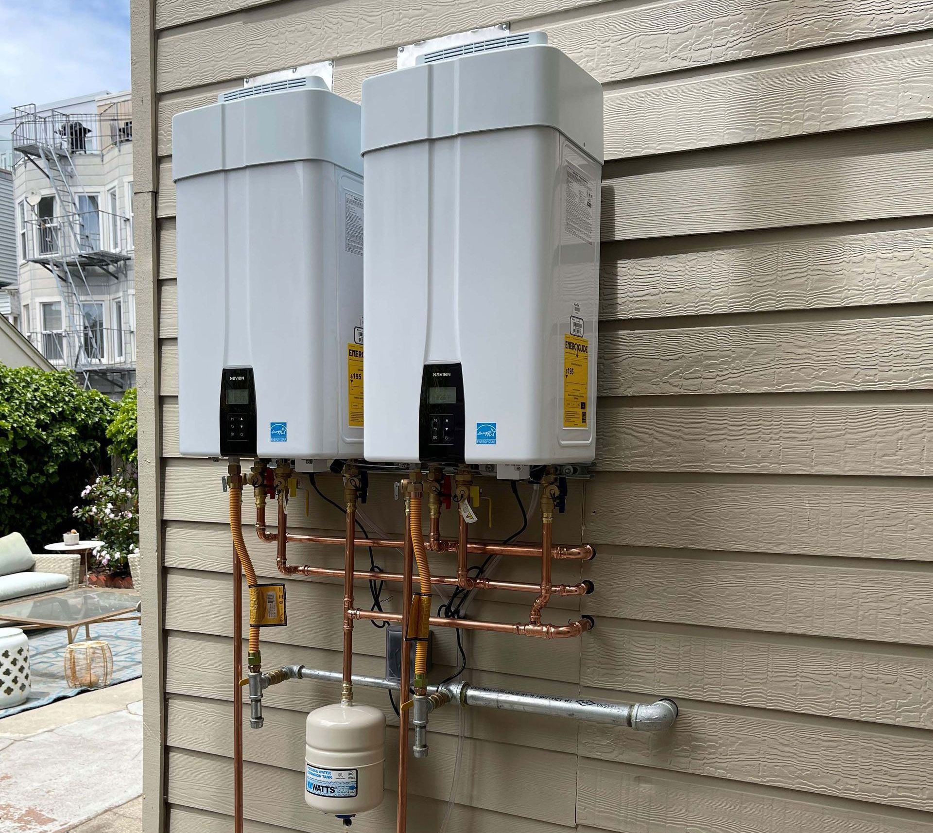 Two white tankless water heaters mounted on a beige, wood-paneled exterior wall with copper pipes.