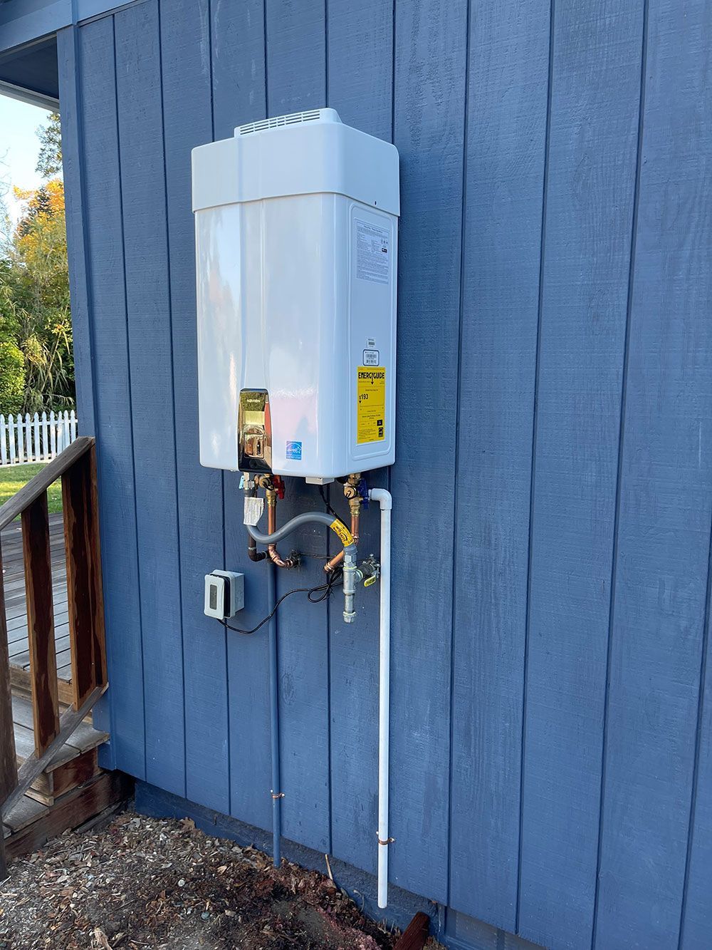A white tankless water heater mounted on a blue siding wall outdoors, with exposed pipes and a gas line.