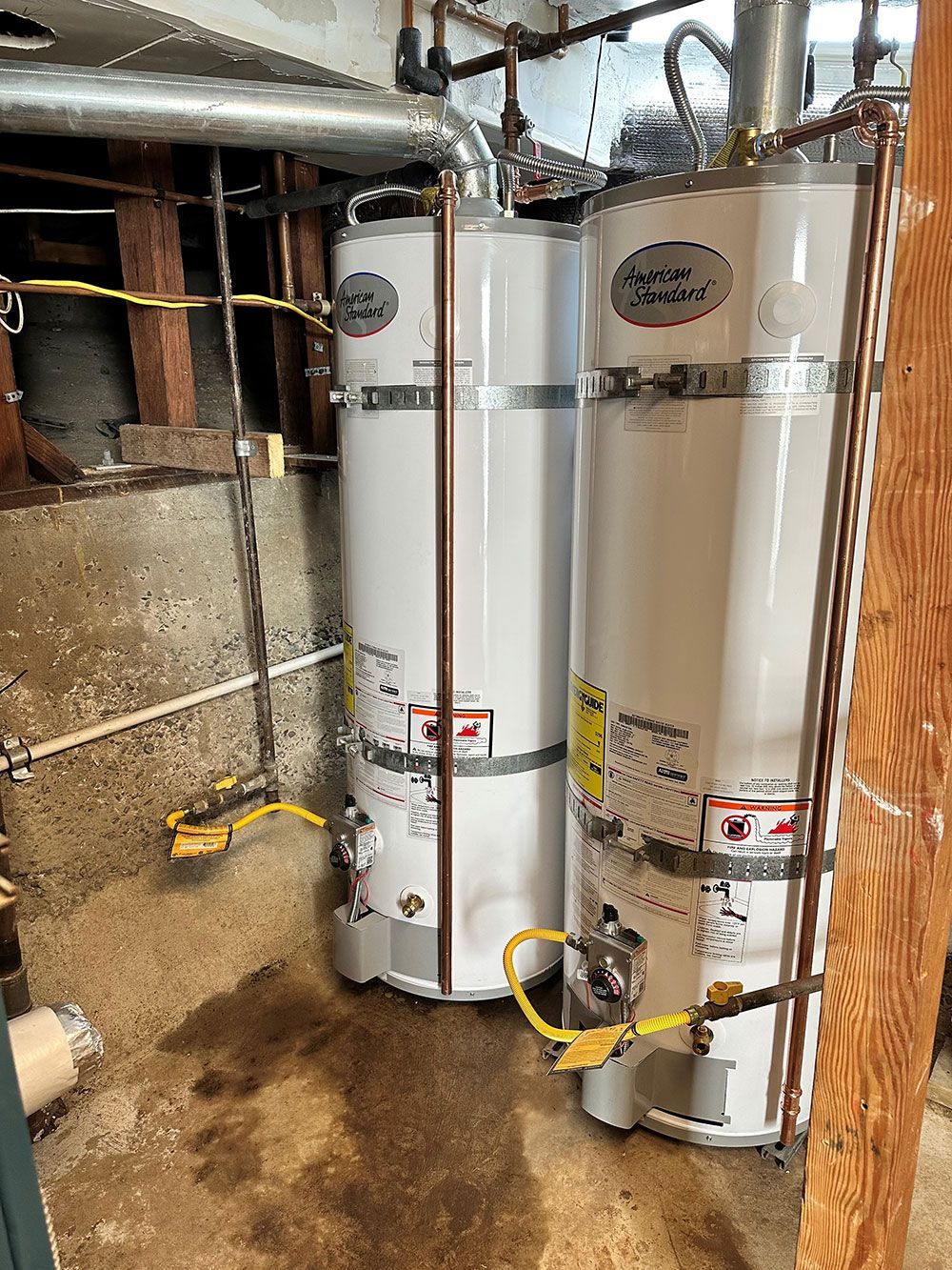 Two tall, white water heaters with copper pipes in a dimly lit basement. Yellow gas lines are connected to the heaters.