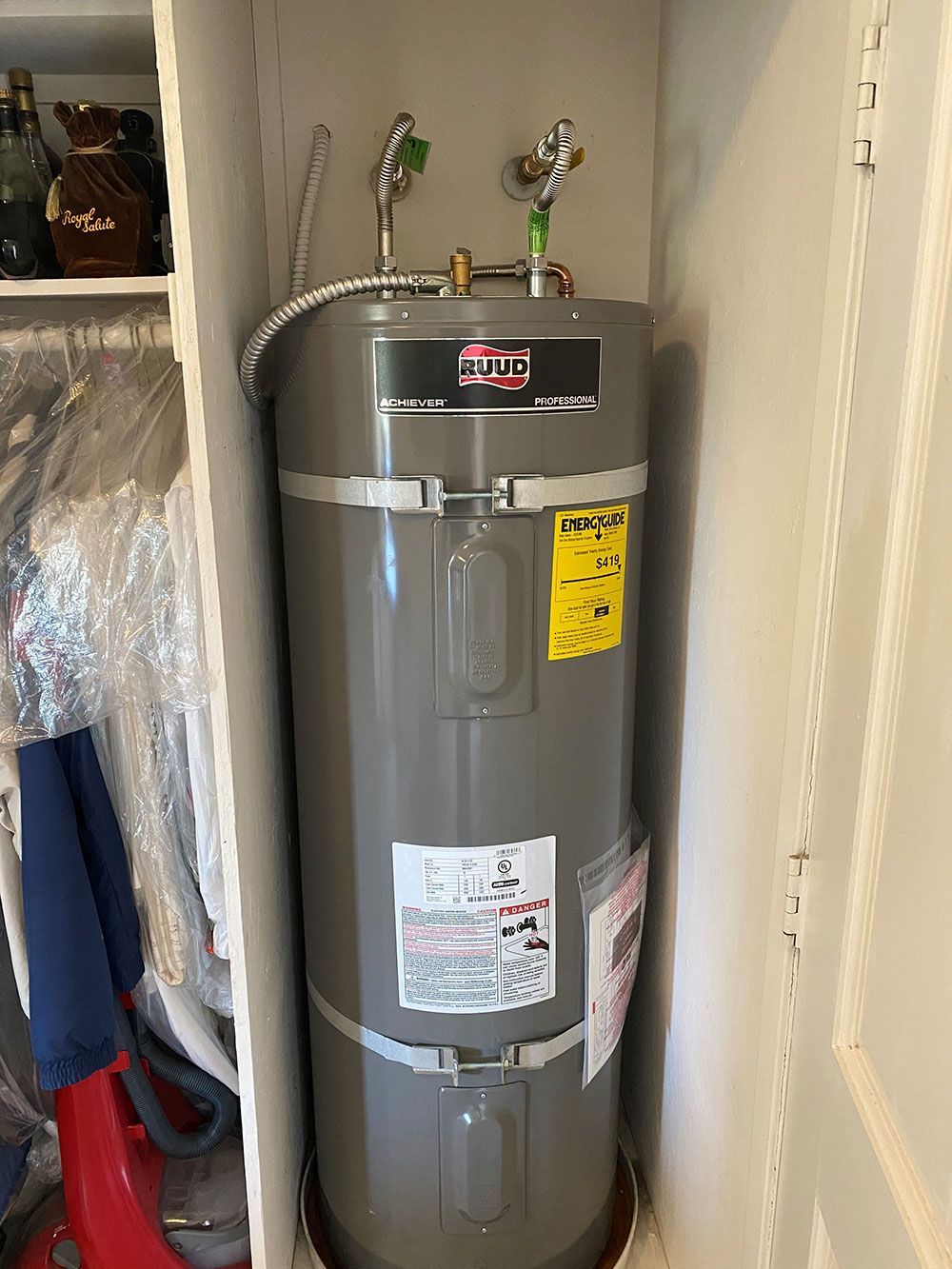 A tall, gray water heater stands in a closet, flanked by shelves with items like bottles and clothing.