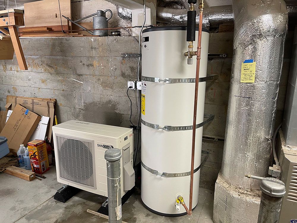 A heat pump water heater in a basement setting. The unit is next to a ventilation duct and an air conditioner.