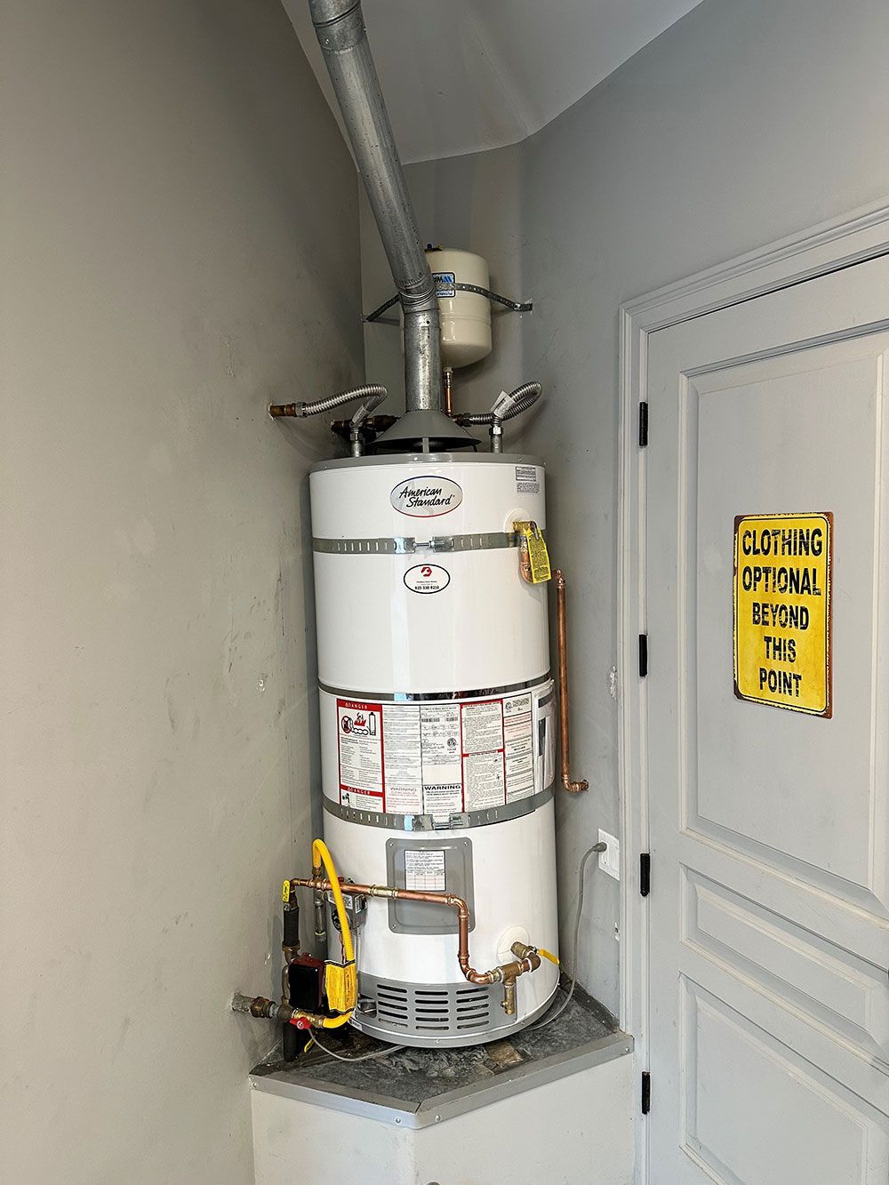 A natural gas water heater in a corner of a room. The heater is white with copper piping and connected to a vent. A door and caution sign are nearby.