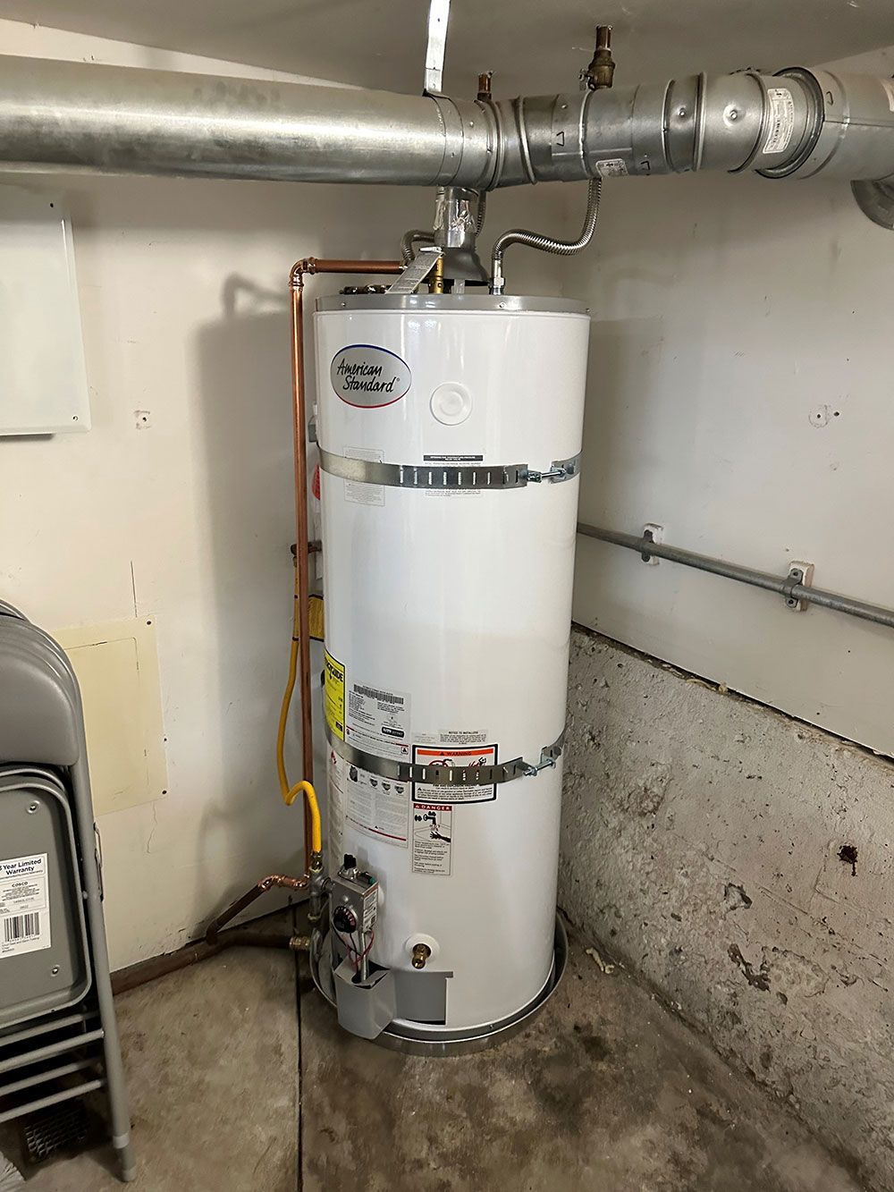 A white water heater in a garage with pipes and a yellow gas line. The setting is a dimly lit, indoor space.
