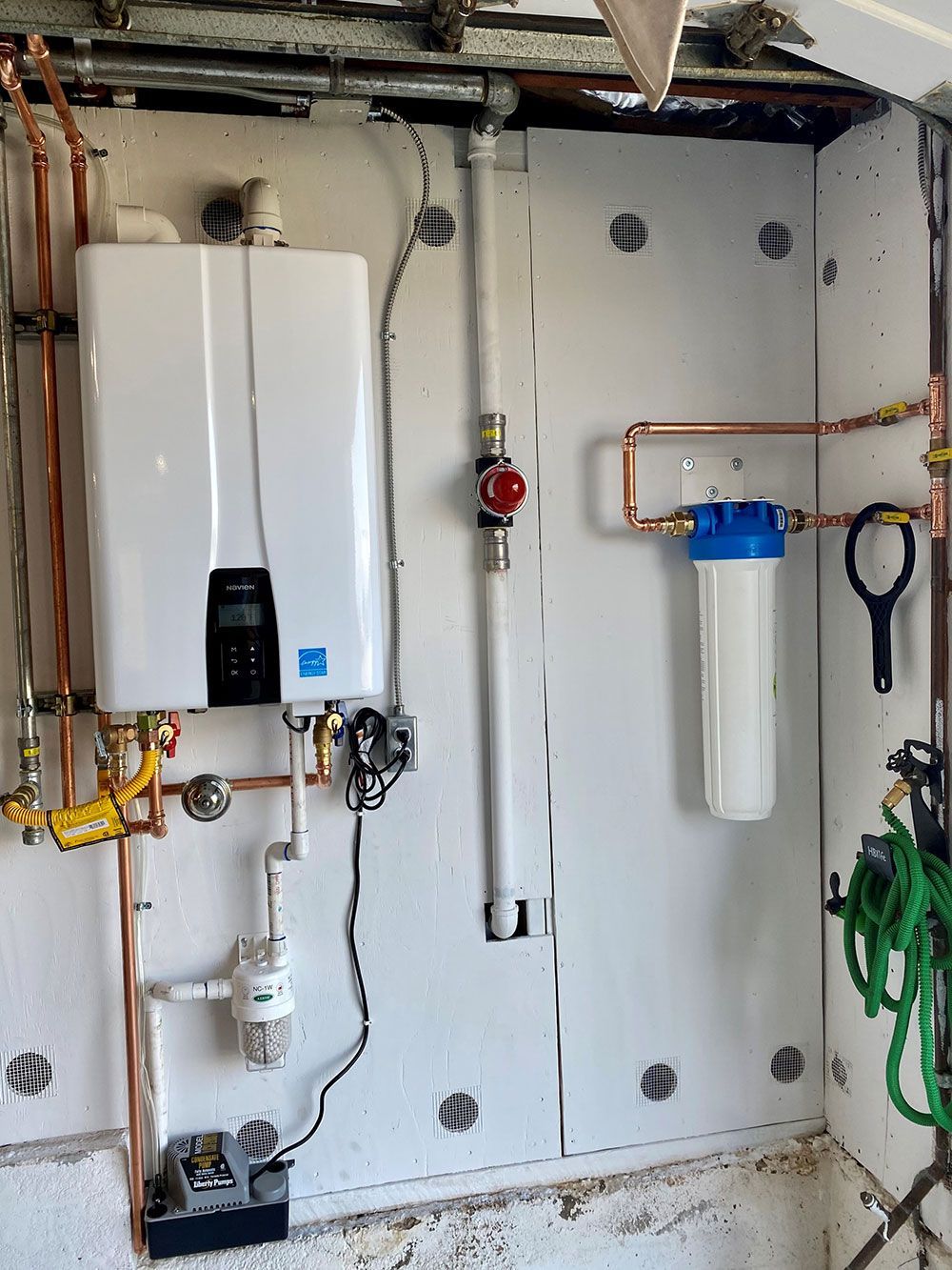 White tankless water heater and water filter mounted on a white wall. Copper pipes and a gas valve are also visible.