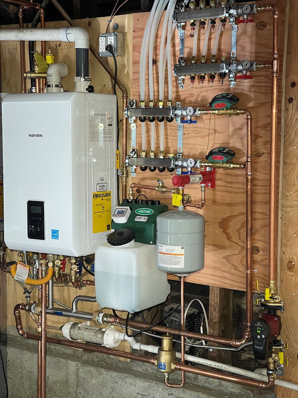 A wall-mounted gas boiler system with copper pipes and various components for heating, installed indoors against a wooden wall.