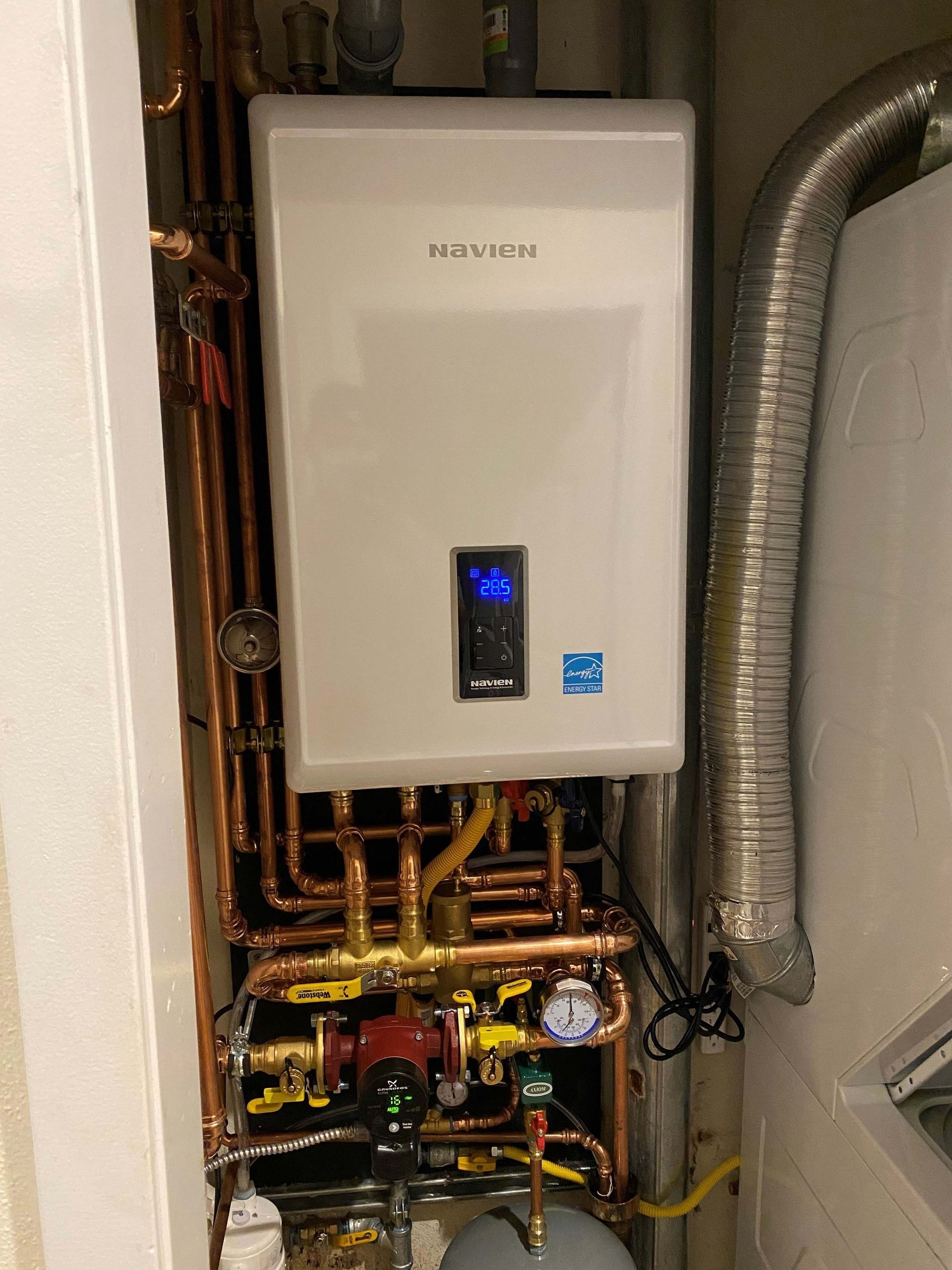 A white tankless water heater with blue digital display is mounted on a wall. Copper pipes and a vent are connected, and the setting appears to be a utility room.