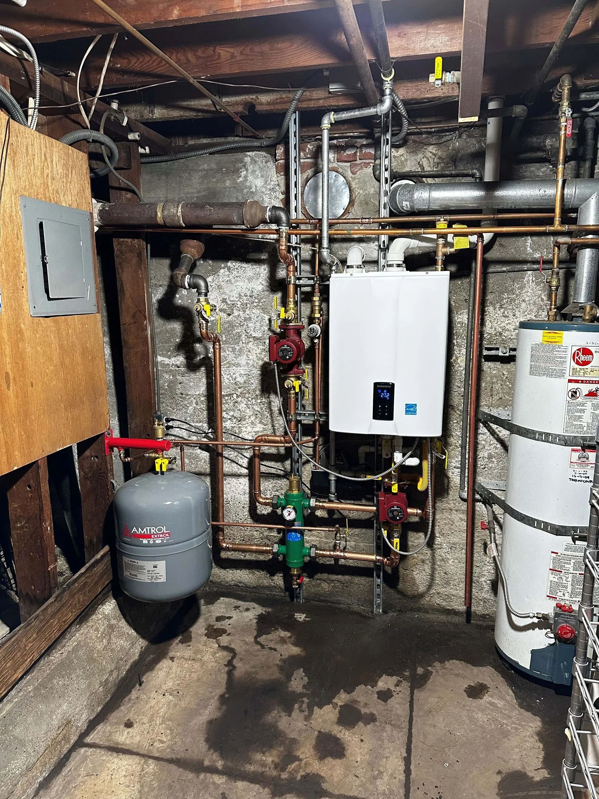 Basement with heating system components: water heater, boiler, pumps, and pipes against a concrete wall. A gray expansion tank is to the left.