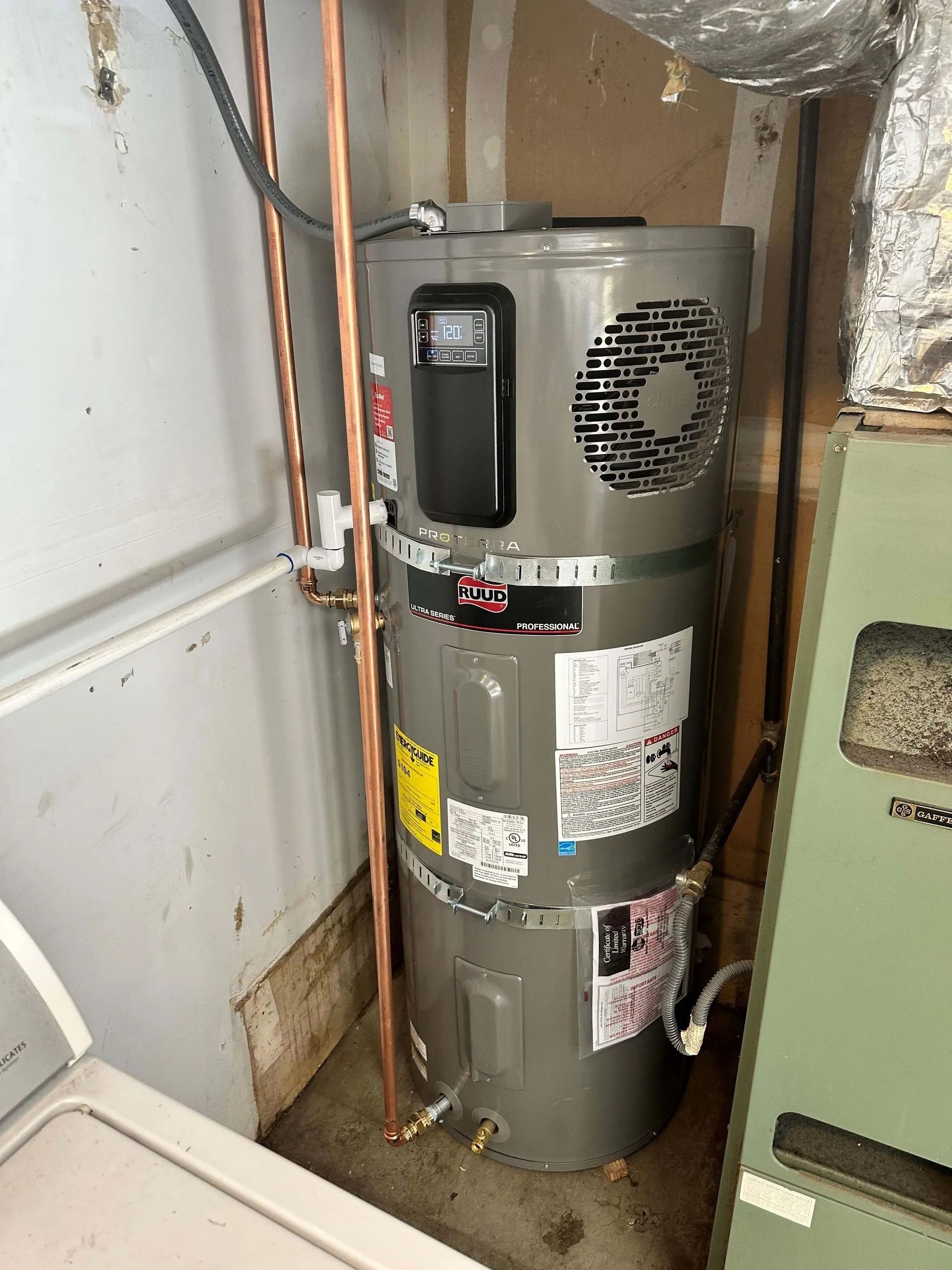 A tall, gray heat pump water heater installed in a utility room. Copper pipes run alongside it, and a green appliance is to the right.