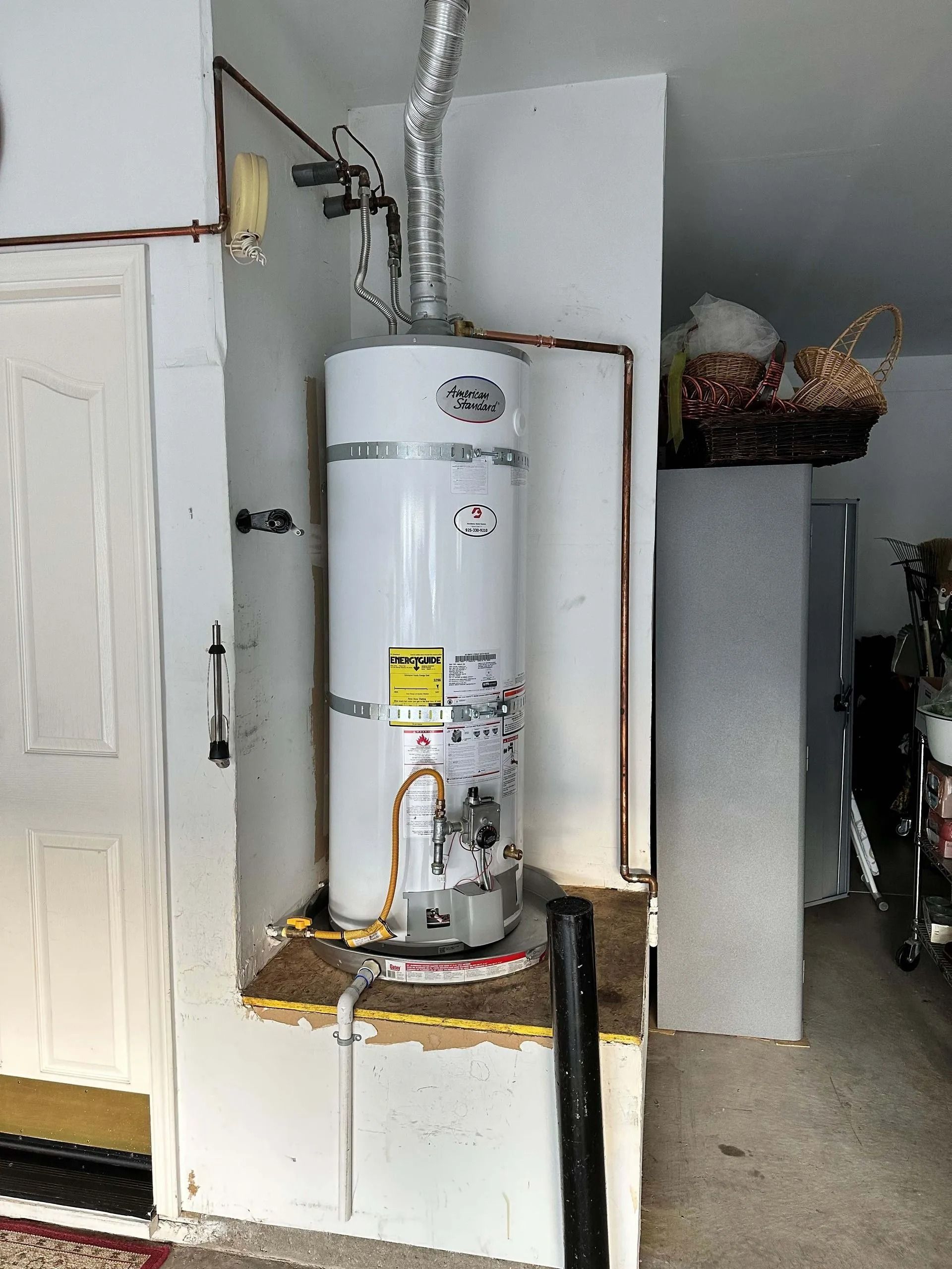 White water heater in a garage, with a silver vent pipe. Copper pipes and a black pipe are visible.