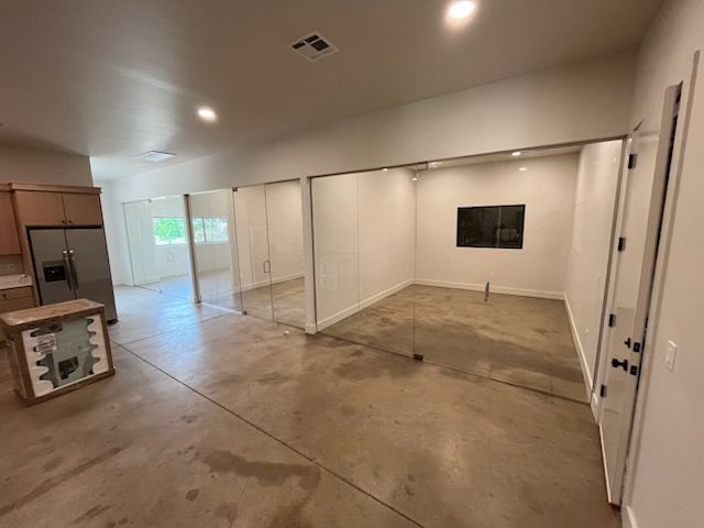 Empty open-plan room with concrete floor, white walls, and recessed ceiling lights; kitchen area at left.