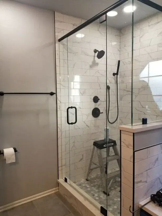 Modern bathroom with glass walk-in shower, marble tile, black fixtures, and a small stool inside.
