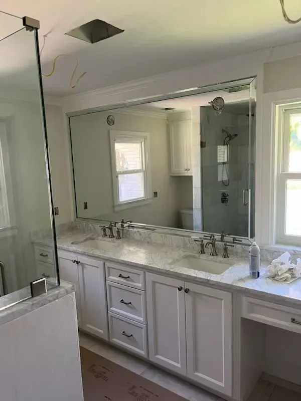 Bright bathroom vanity with large mirror, double sinks, white cabinets, and a glass shower door.