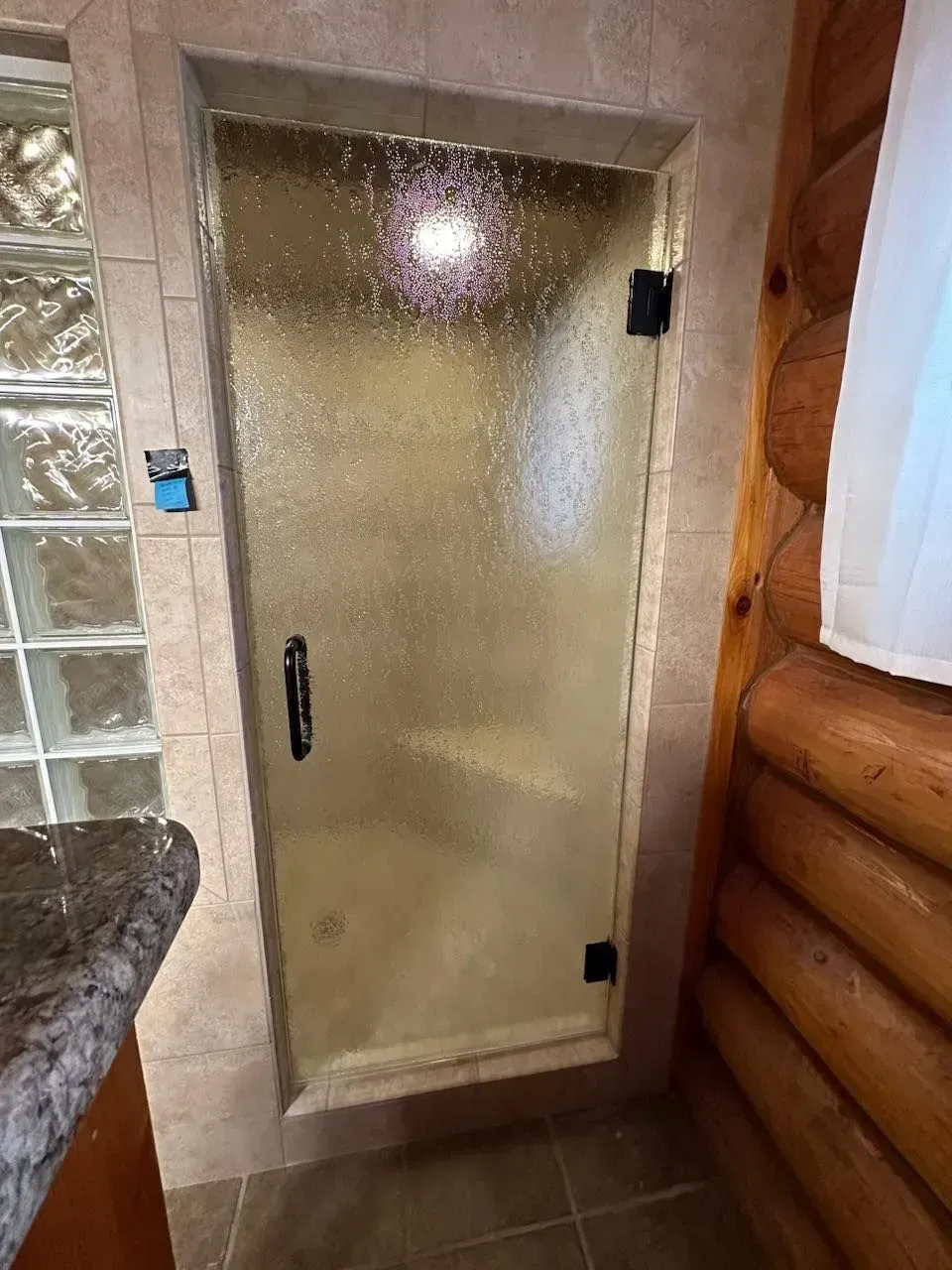 Frosted glass shower door with black handle in a tiled bathroom, next to a log wall and granite counter.