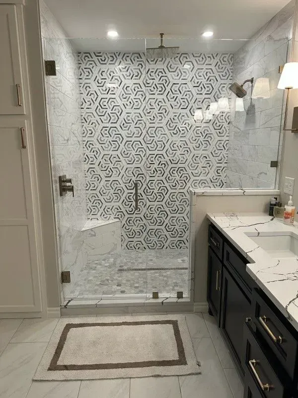 Modern bathroom with patterned shower tile, glass door, dark vanity, and light-colored rug