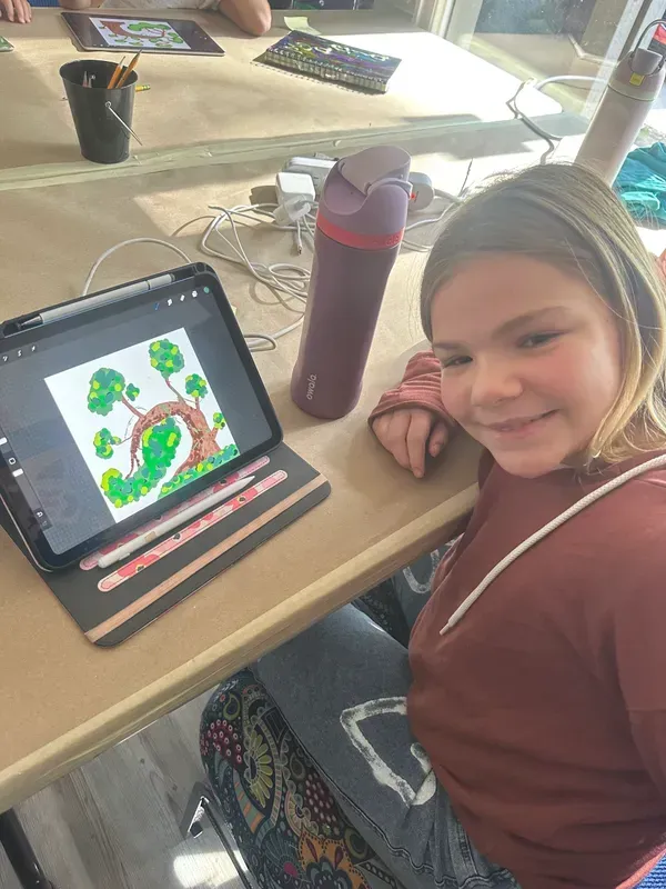 Girl smiling, working on a digital drawing of a tree on a tablet. Brown shirt, sitting at a table.
