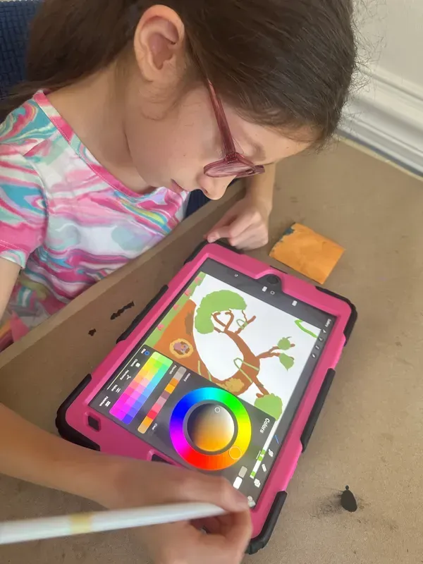 Girl with glasses draws on a pink tablet, selecting colors with a stylus. Tree drawing visible.