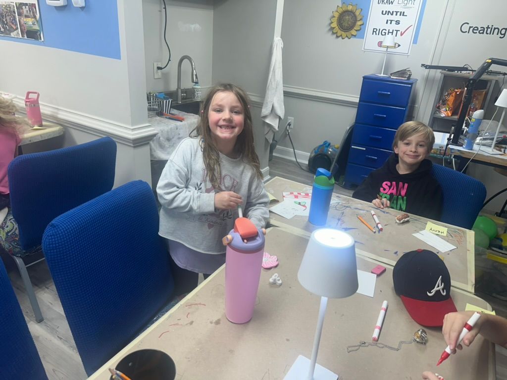 Two kids smiling at a table, likely crafting in a room with blue chairs, water bottles, and a lamp.
