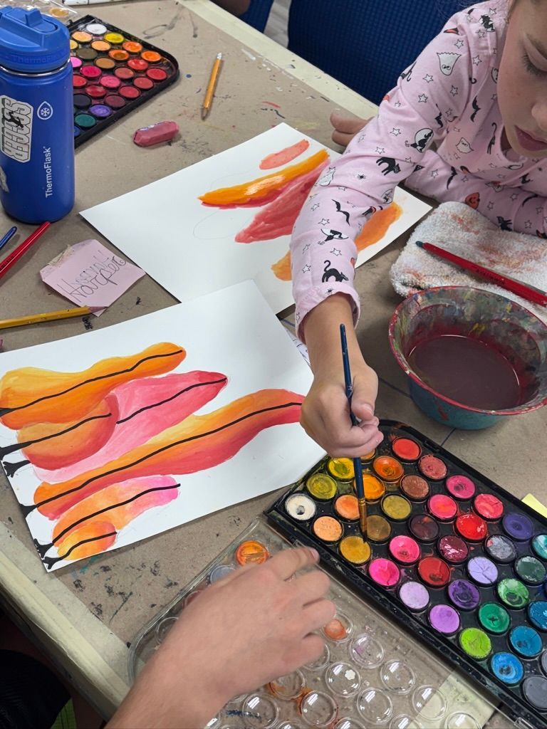Child painting with watercolors at a table. Orange and red brush strokes on paper, paints, and a water cup are visible.