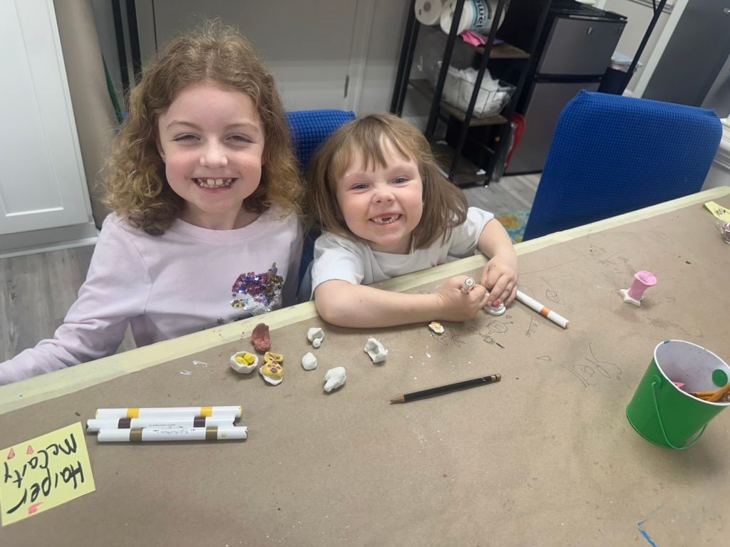 Two smiling children at a table, crafting with clay.  They sit in a room with craft supplies.