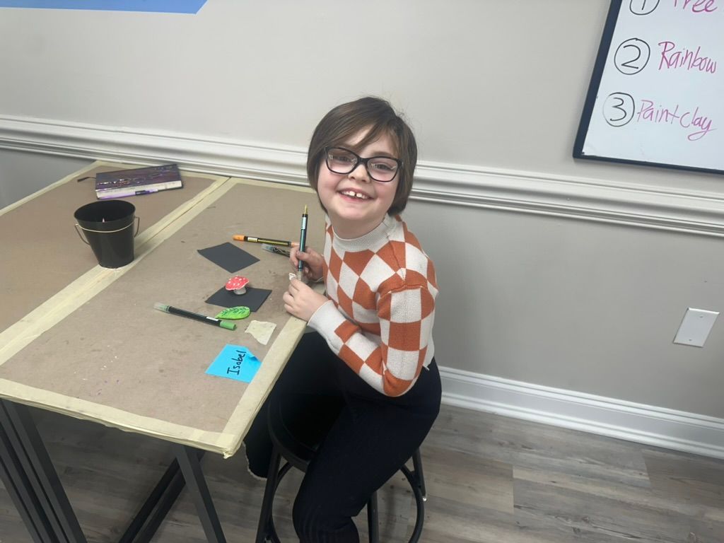 A person with glasses smiles while sitting at a table with art supplies.