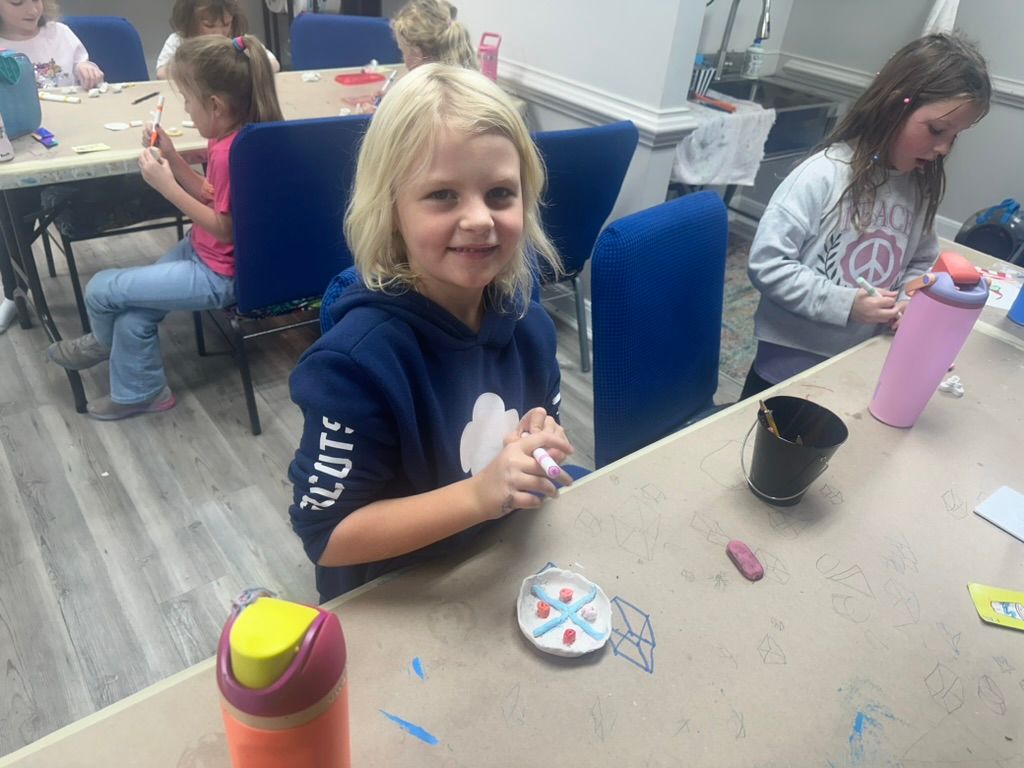 Girl smiling at the camera at a craft table, working on a project with other children.