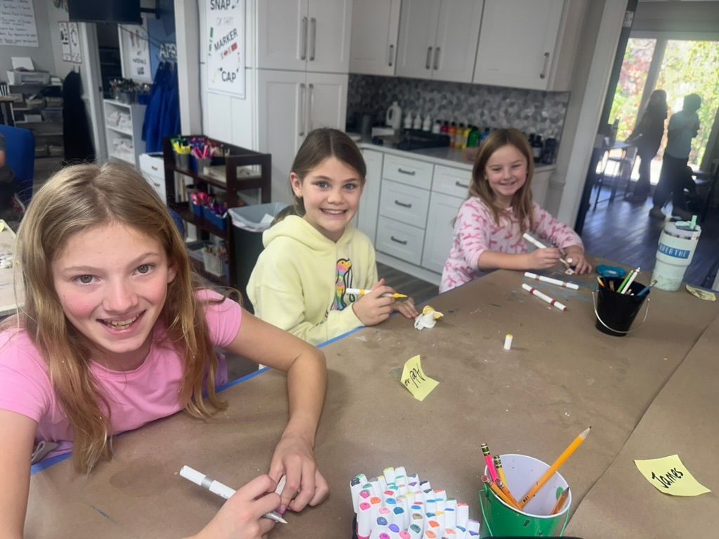 Three girls smiling and crafting at a table in a brightly lit room.