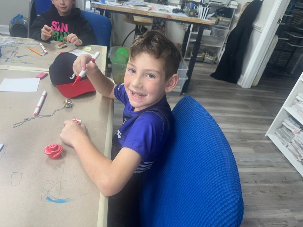 Boy smiling, holding a marker at a table, hat and supplies present. Another person in background.