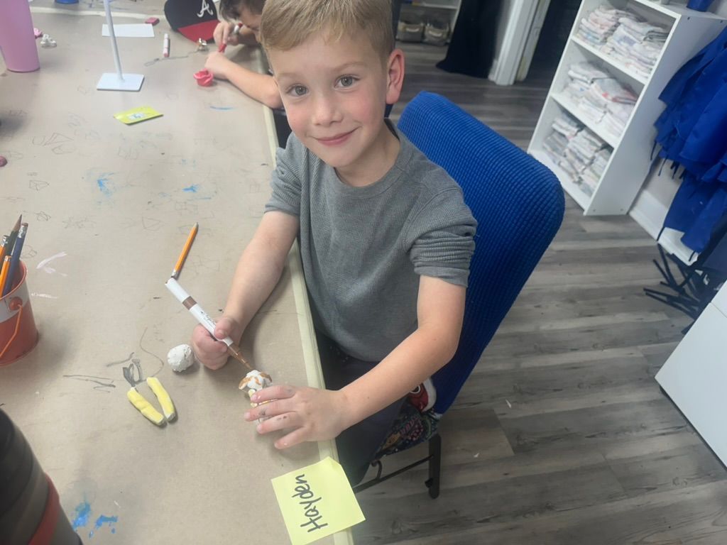 Boy at a table smiles, holding a pen and crafting supplies. Yellow name tag reads 