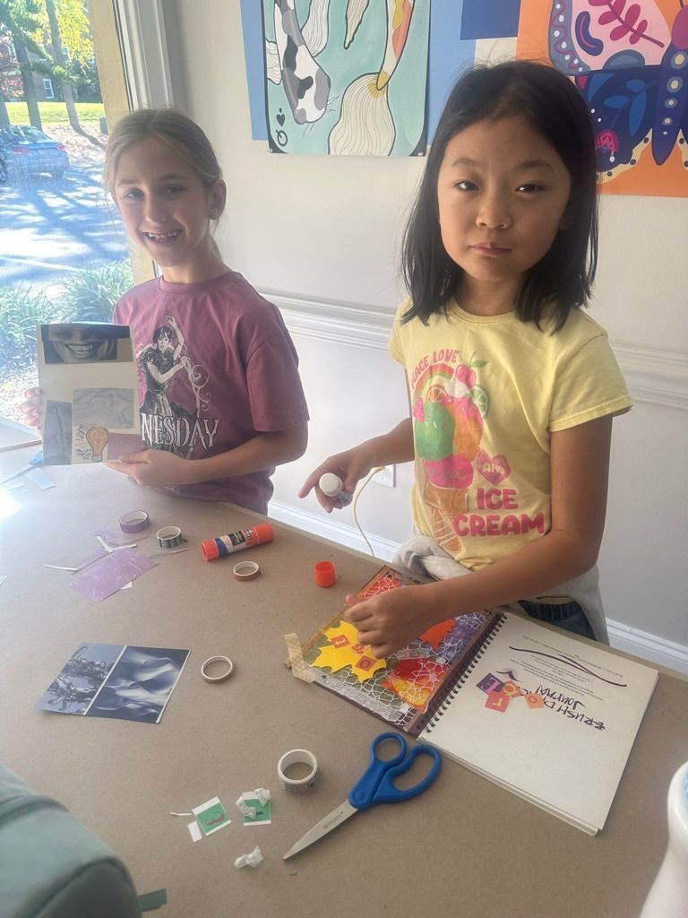 Two girls crafting at a table with paper and supplies. One smiles, the other looks at the camera.