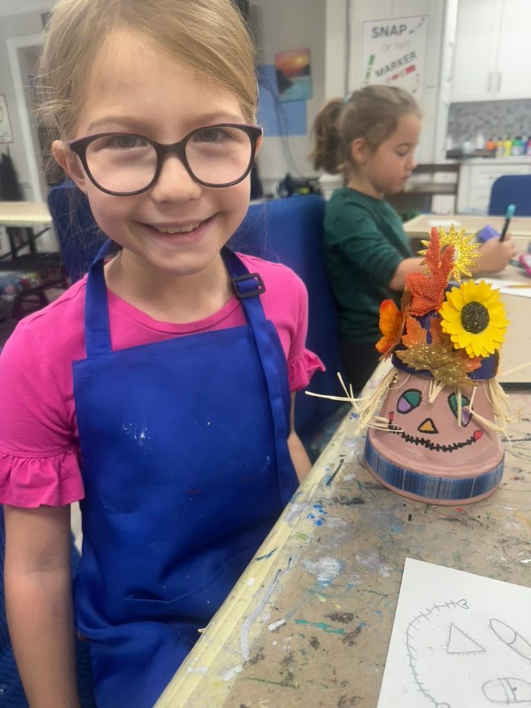 Girl in glasses smiles, wearing an apron, next to a pumpkin-themed craft project. Another girl in the background.