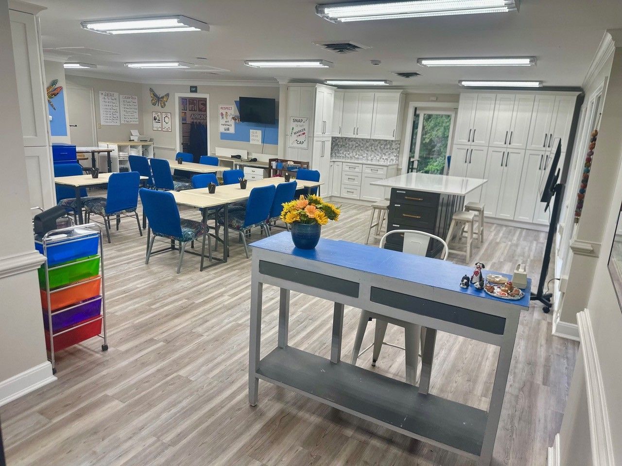 Bright classroom with blue chairs, tables, a gray cart, and colorful bins under fluorescent lights