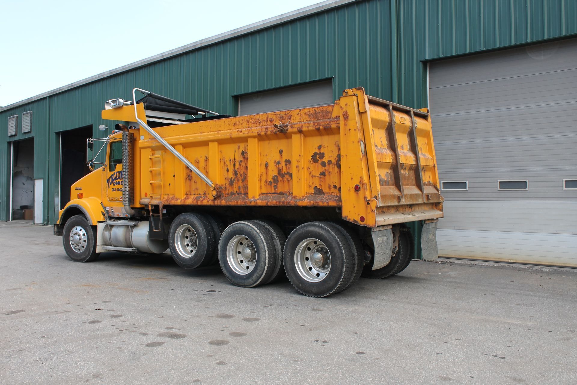A yellow dump truck is parked in front of a green building.