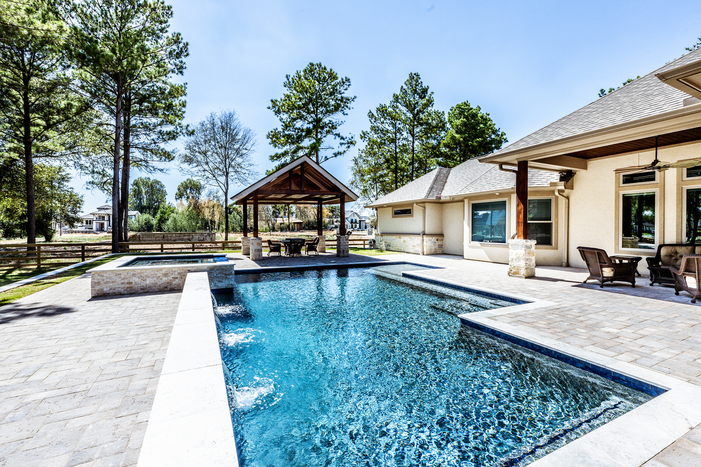Swimming pool and spa in a backyard with a gazebo, trees, and a beige house.