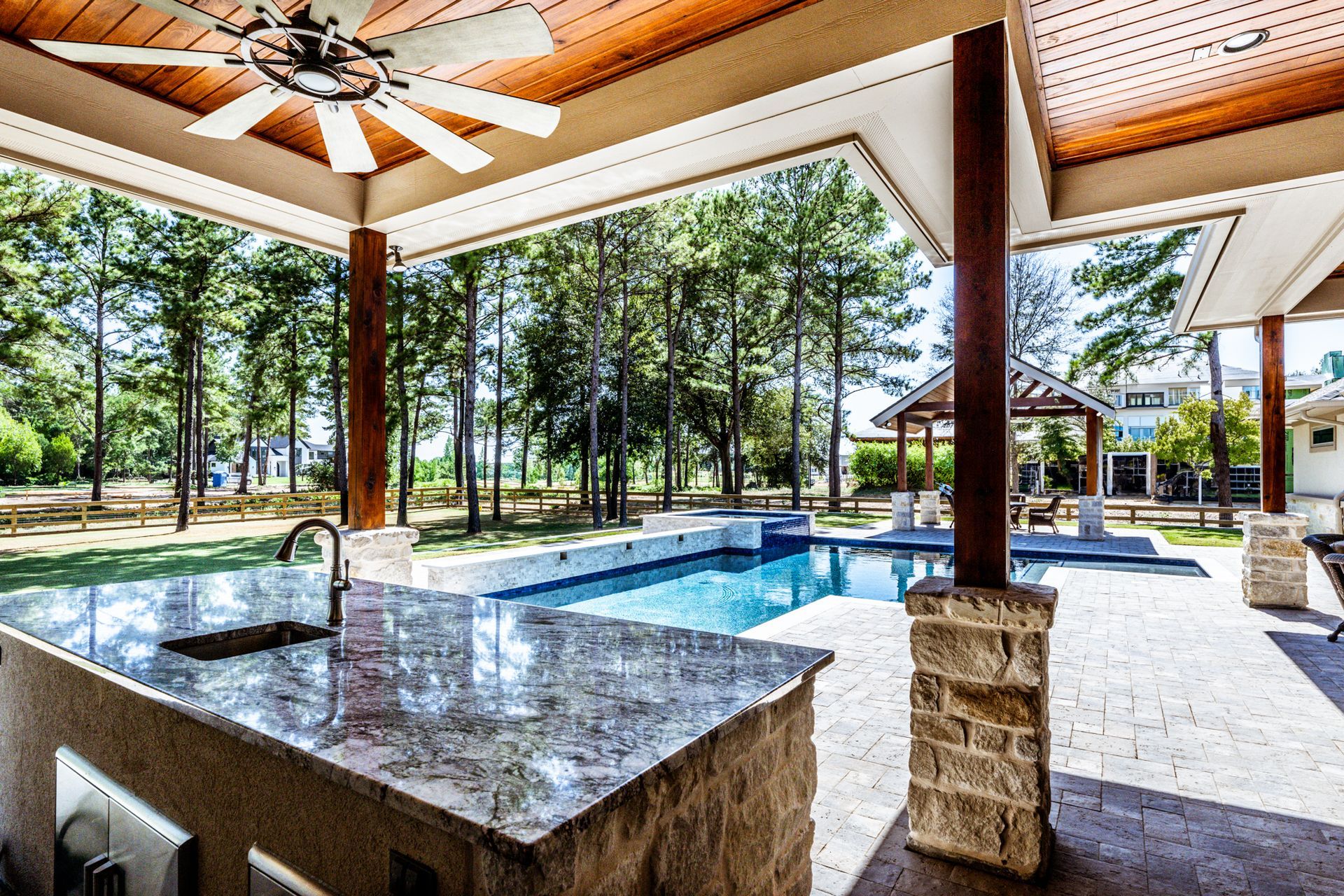 Outdoor kitchen with a granite countertop, pool, and trees in the background.