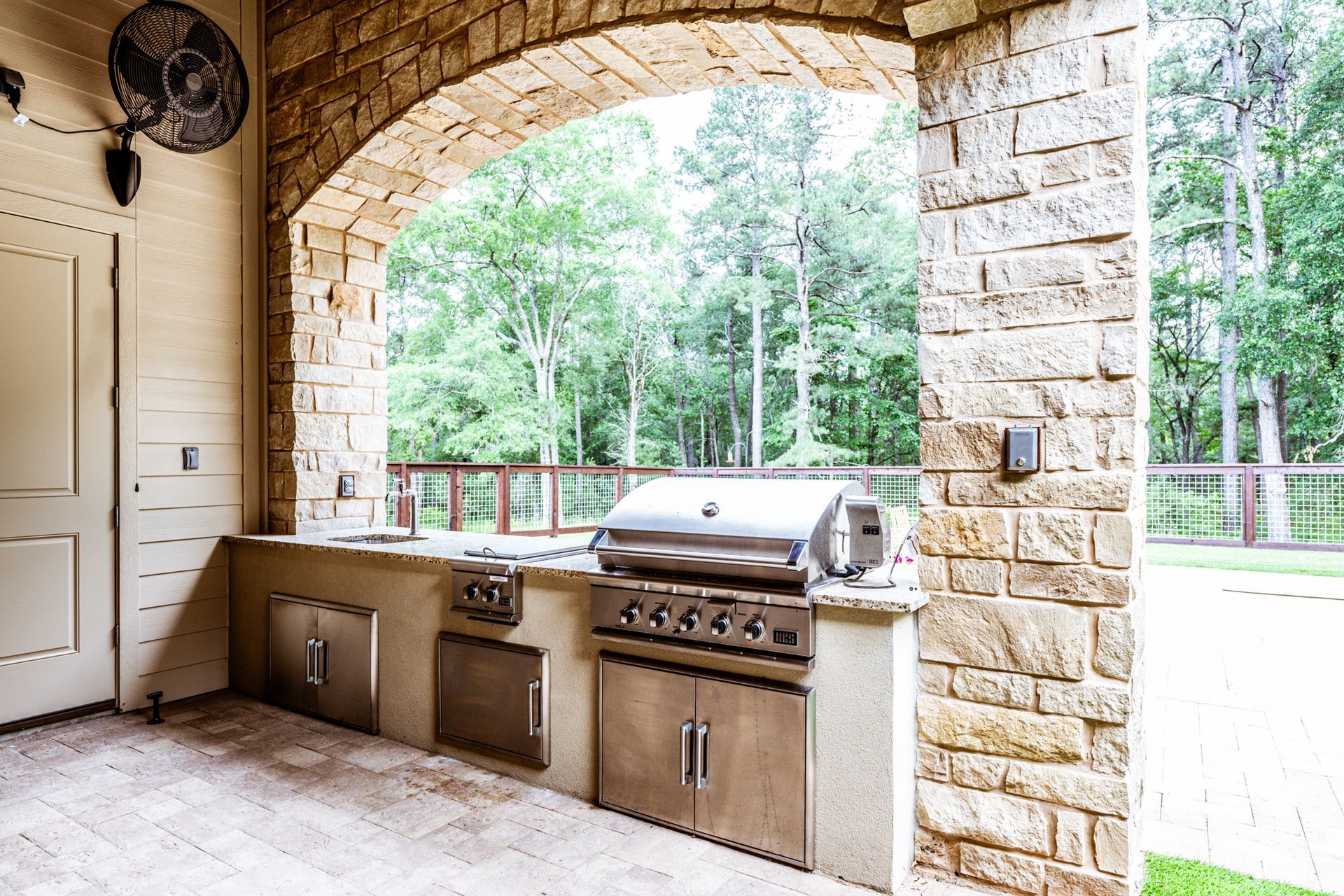 Outdoor kitchen with stone archway, stainless steel grill and cabinets, overlooking a wooded area.