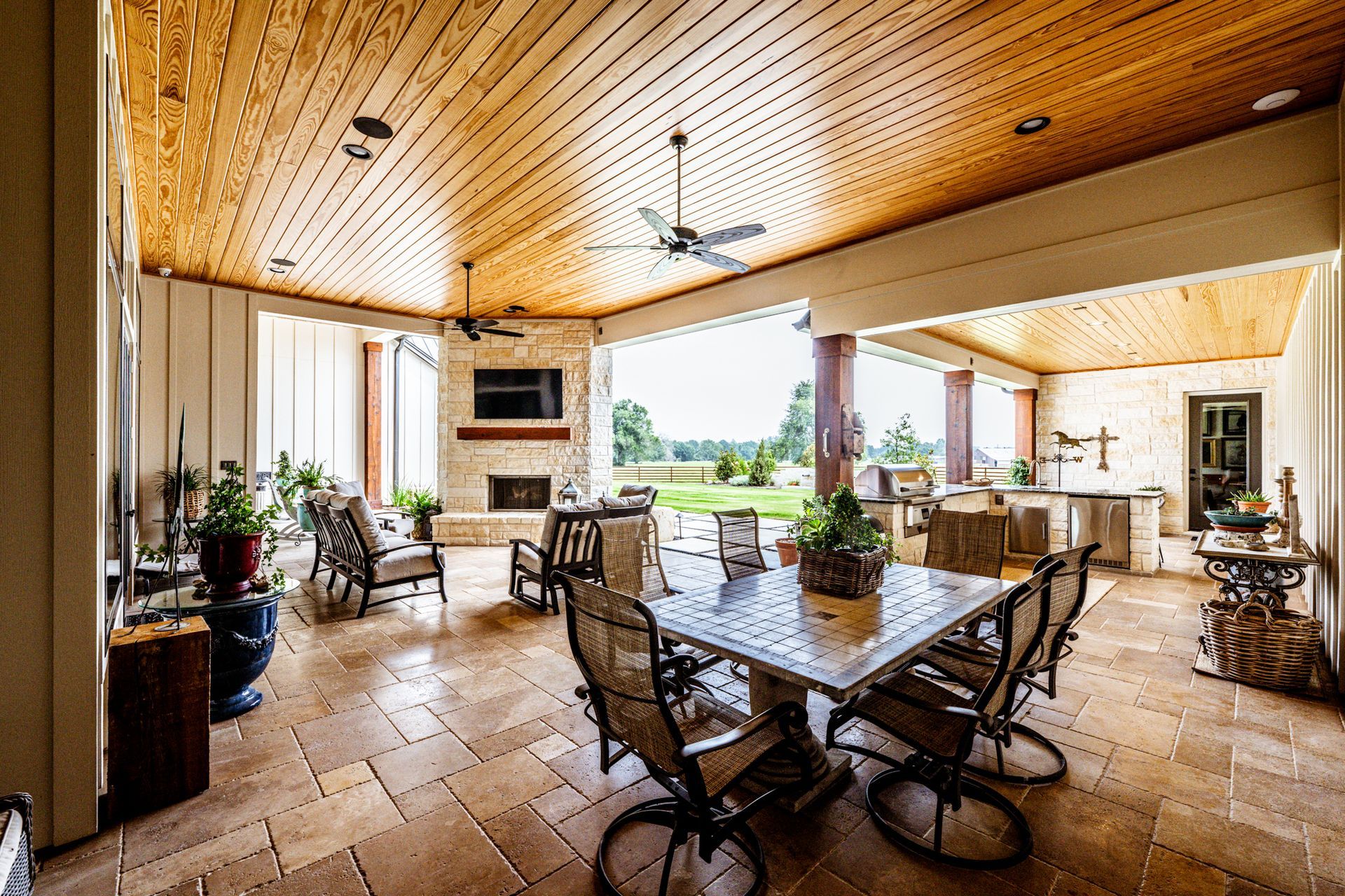 Covered patio with dining table, fireplace, and outdoor kitchen overlooking a green field.