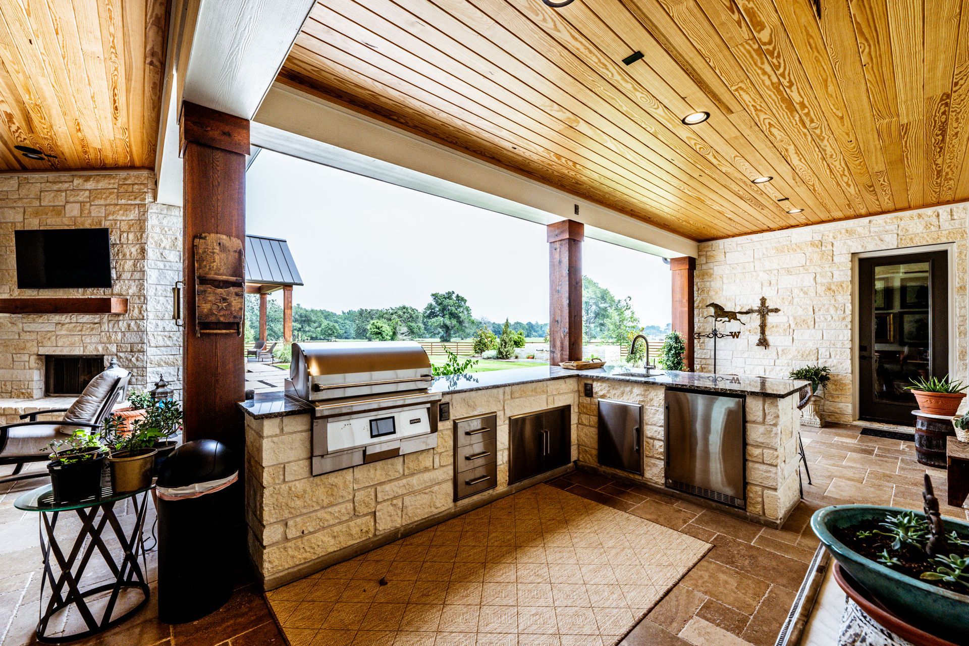 Outdoor kitchen with grill, counter, and stainless steel appliances under a wood ceiling with a view of a green landscape.