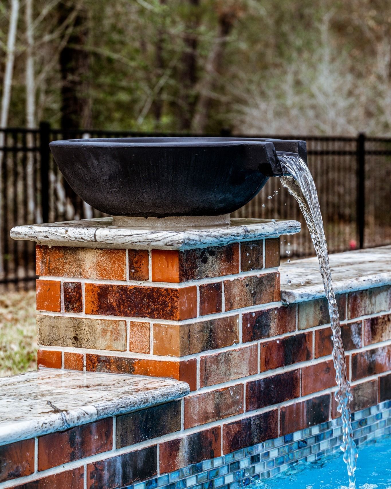 A water fountain is sitting on top of a brick wall next to a swimming pool.