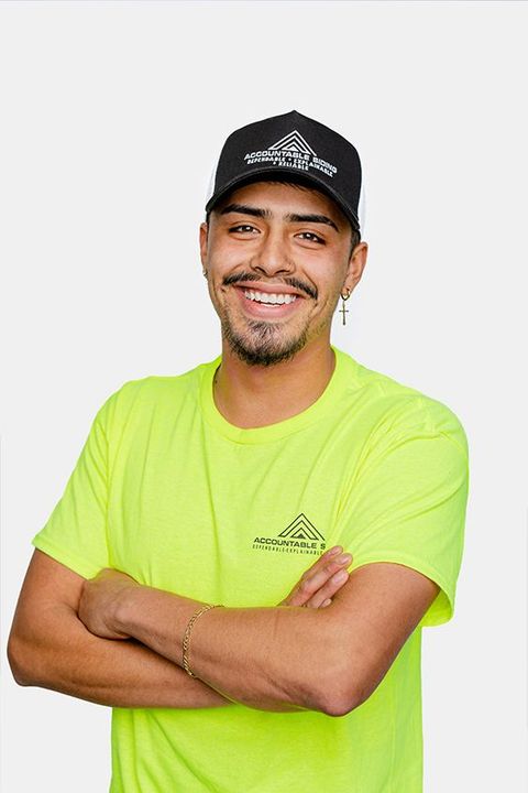 A man wearing a yellow shirt and a black hat is standing with his arms crossed and smiling.