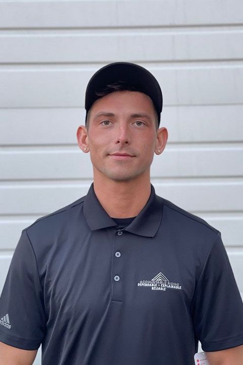 A man wearing a black polo shirt and a black hat is standing in front of a garage door.