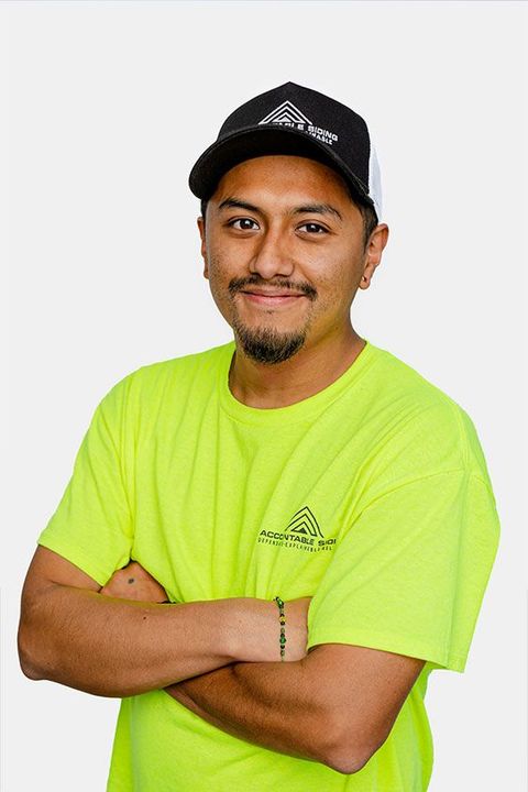 A man wearing a neon yellow shirt and a black hat is standing with his arms crossed.