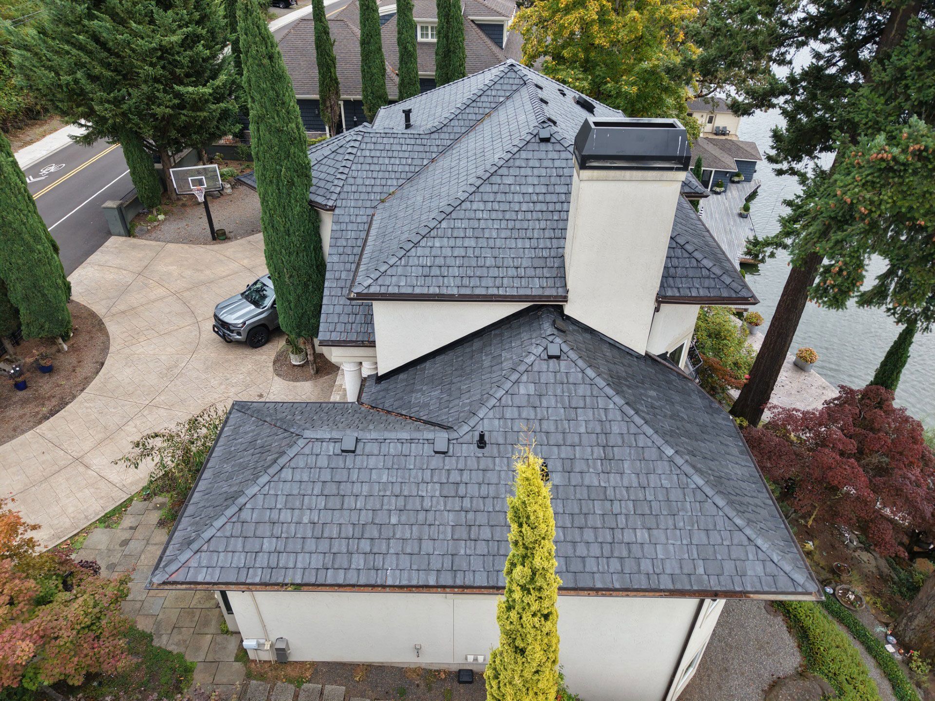 Aerial view of a gray-roofed house with a chimney, surrounded by trees and a driveway.