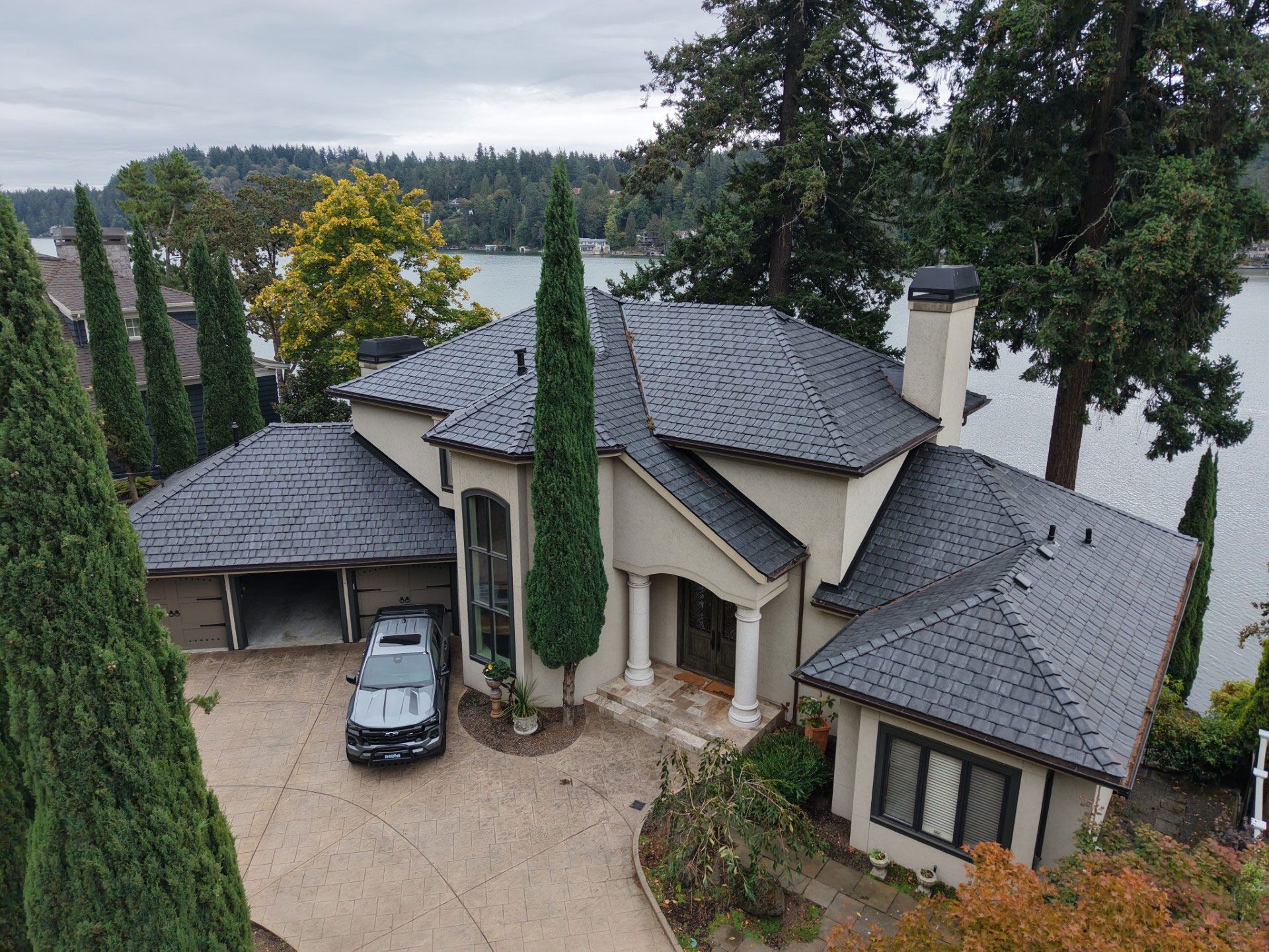 Large beige house with gray roof and parked car, viewed from above near a lake and trees