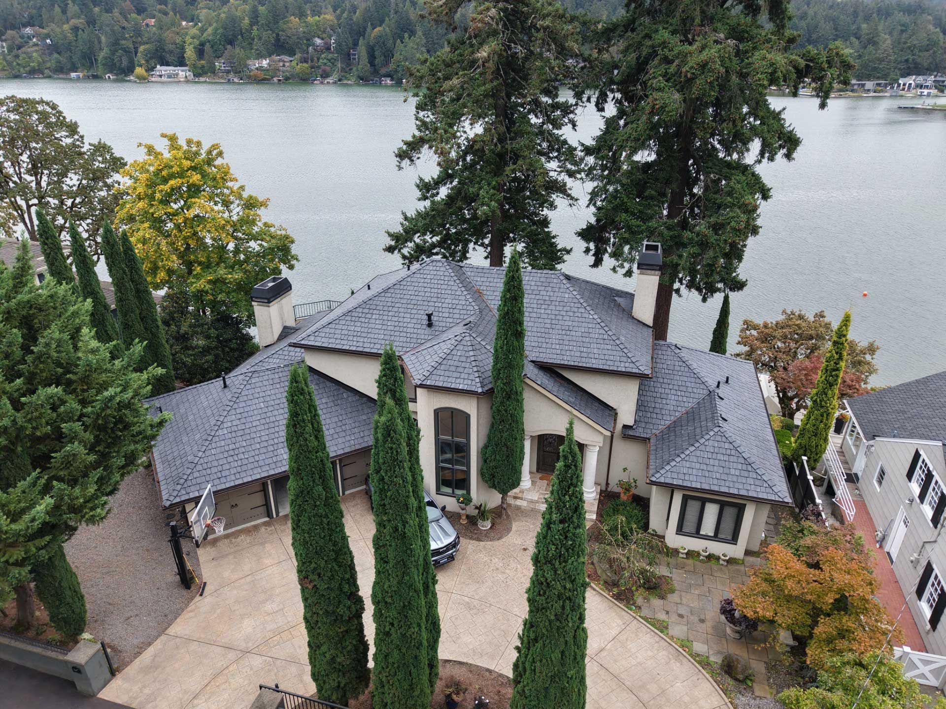 Lakefront house with gray roof, tall evergreens, and a paved driveway