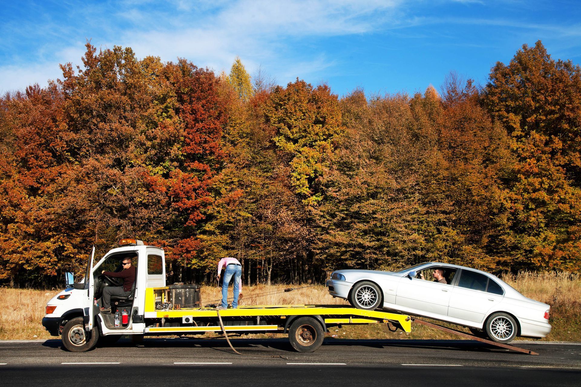 Tow truck with white car on flatbed, two people working. Autumn trees in the background.