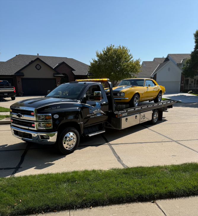 Black tow truck carrying a yellow classic car on a suburban street on a sunny day.
