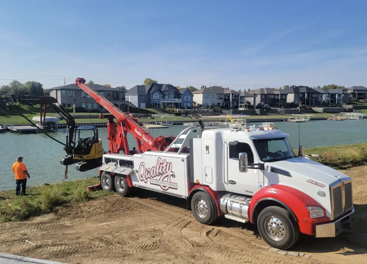 Tow truck with extended crane lifting a small excavator near a body of water with houses in the background.