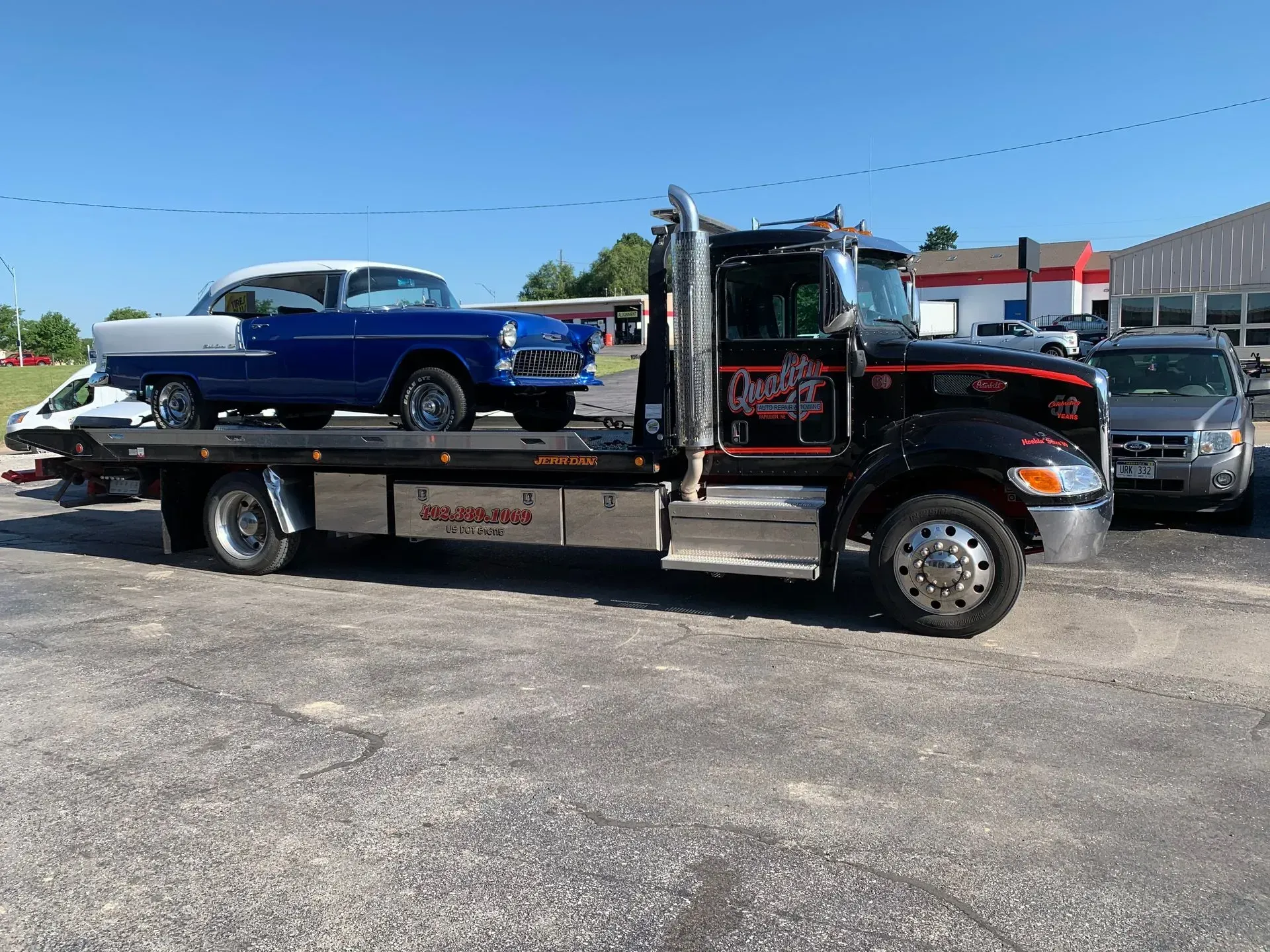 Classic blue and white car loaded on a black tow truck in a parking lot on a sunny day.