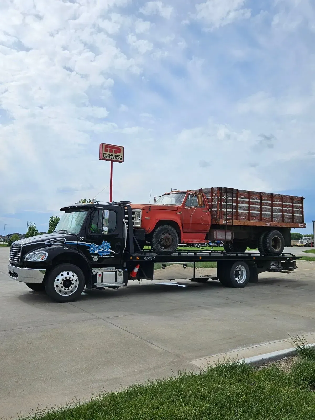 Black tow truck hauling an orange flatbed truck with a load of wood under a cloudy sky.